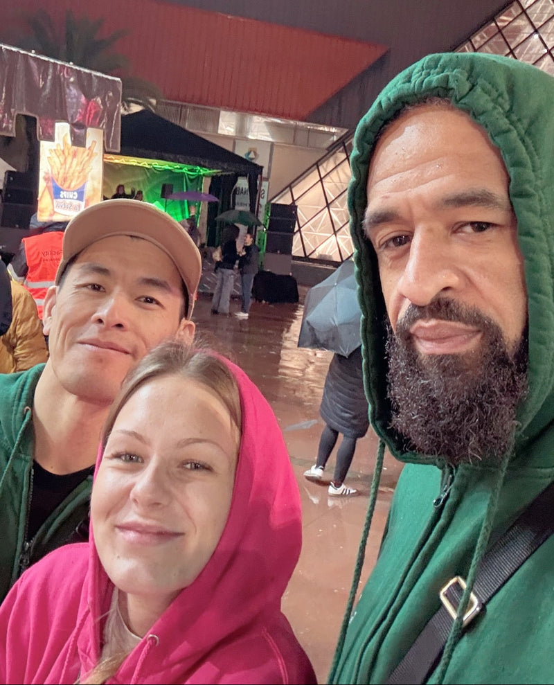 Three people taking a selfie in what looks like an indoor event space or market. The person on the right wears a green hoodie, the person in the middle wears a pink hoodie, and the person on the left wears a tan cap and green shirt.